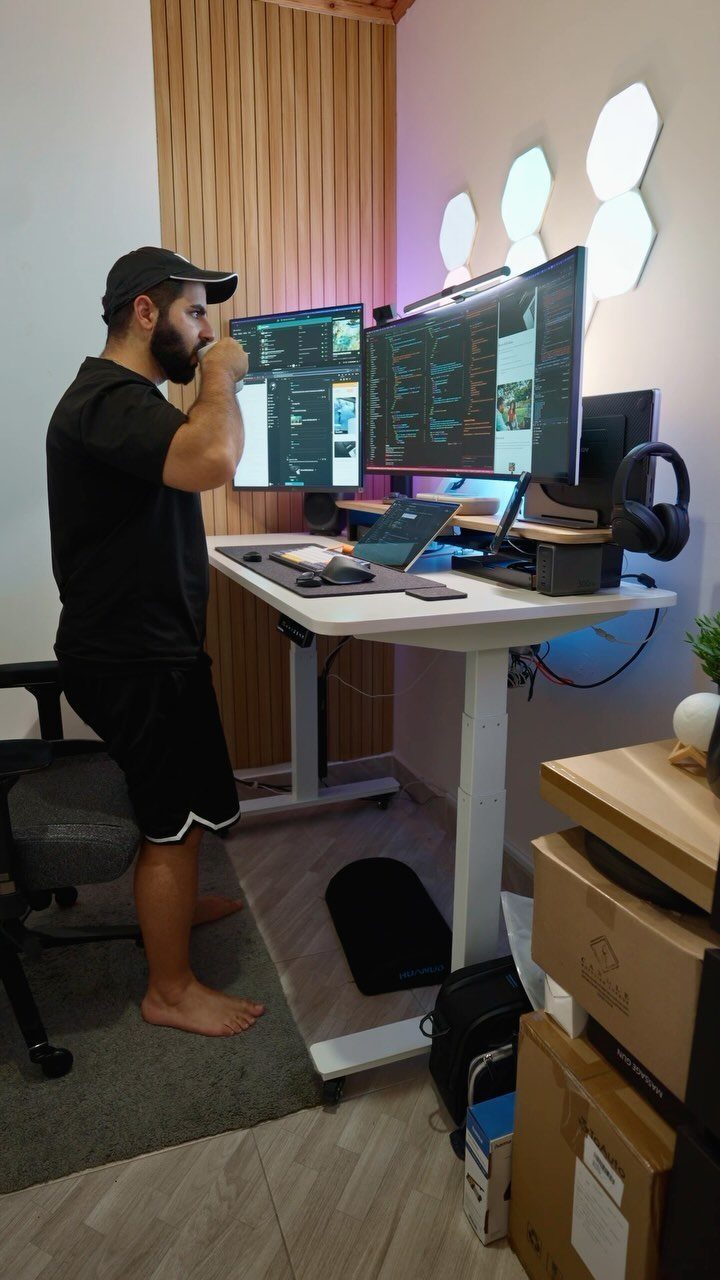 Man standing at a white desk with dual monitors and hexagonal wall lights.