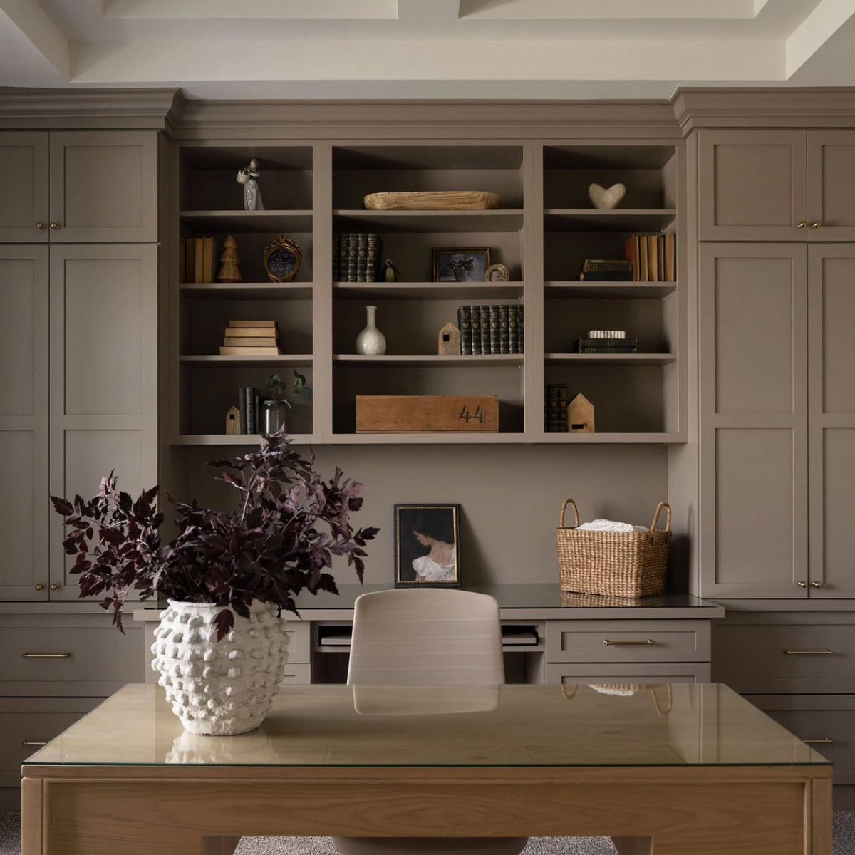 Gray built-in office cabinetry and shelving with a wooden glass-top table and purple foliage.