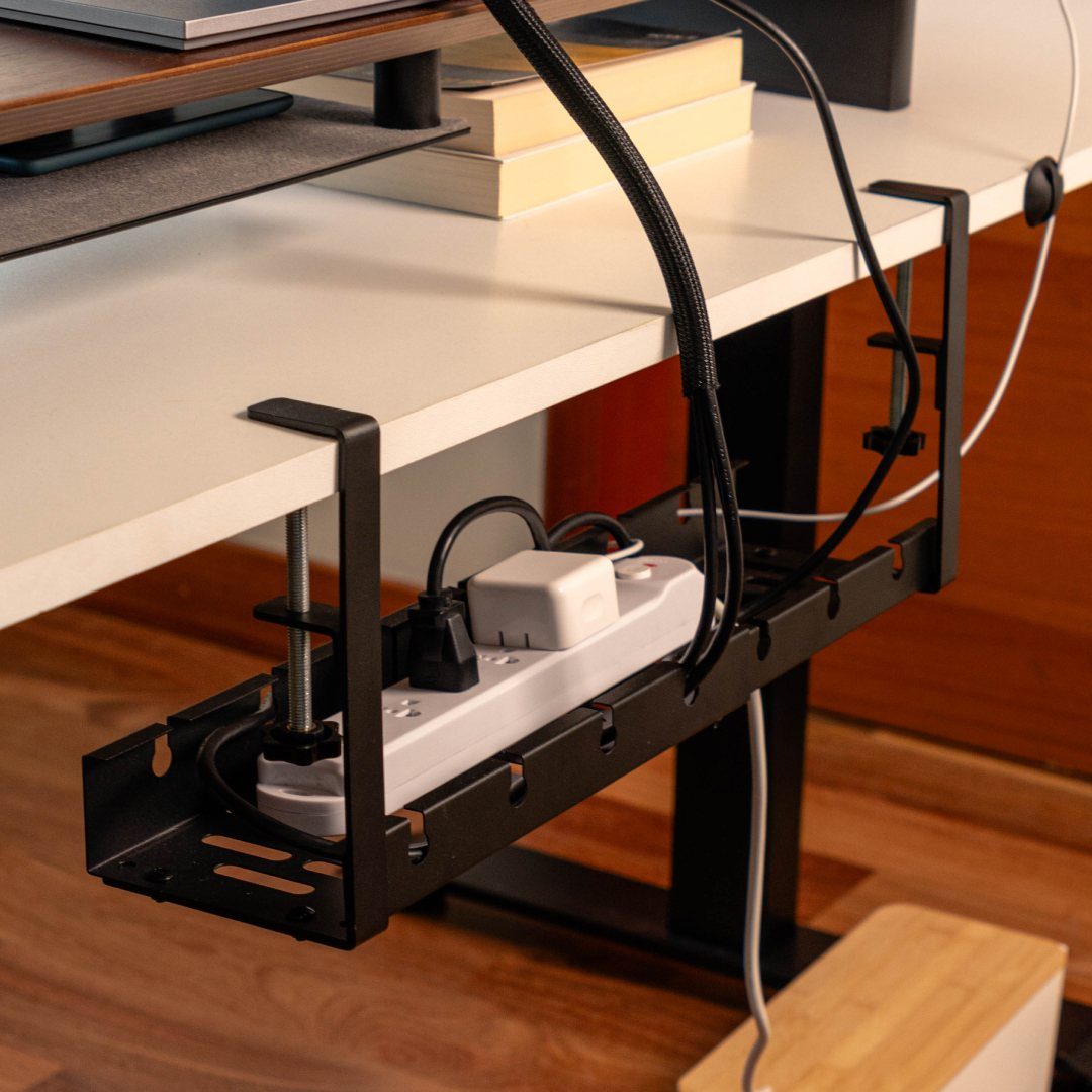 Black metal cable management tray under a white desk holding a power strip.