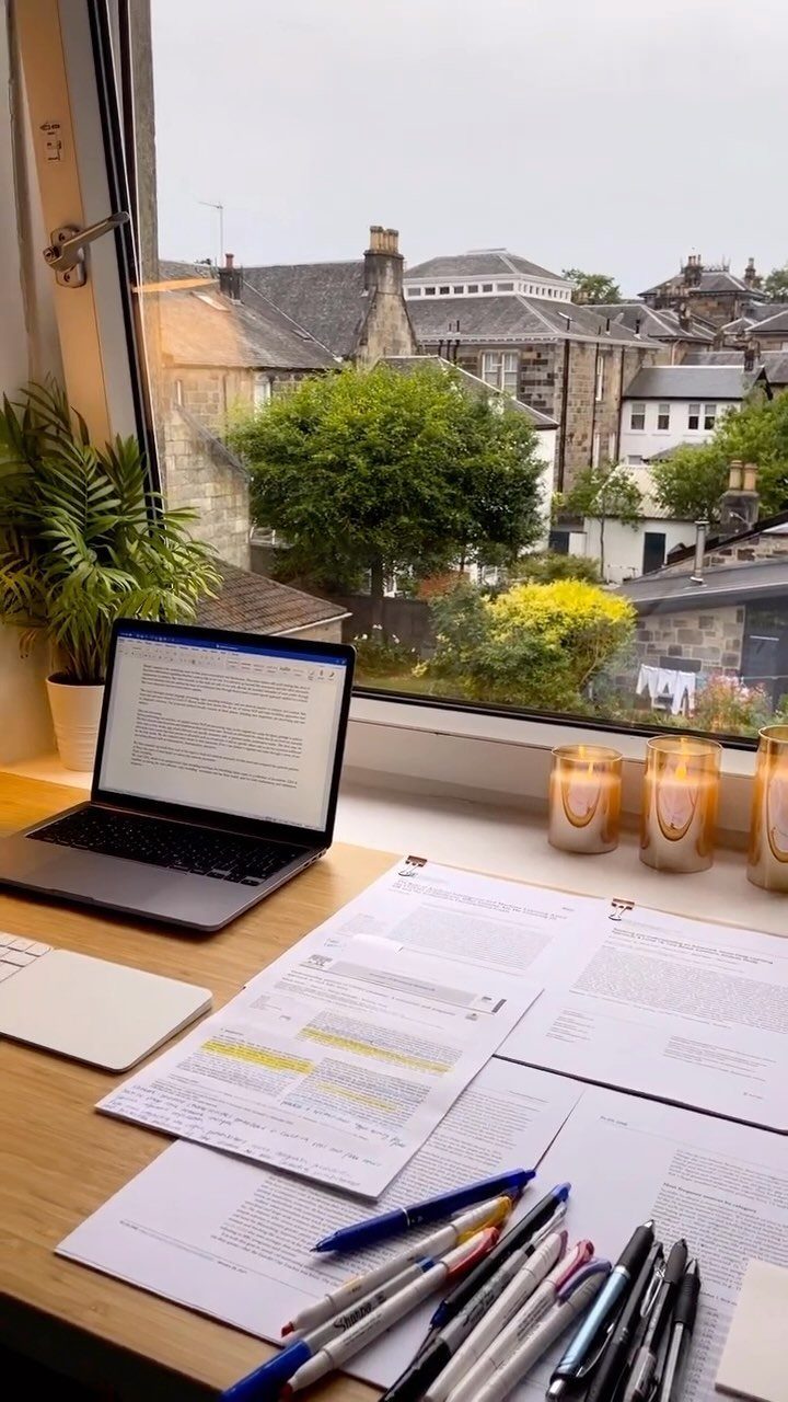 Wooden desk with laptop, highlighted documents, and pens by a city view window.