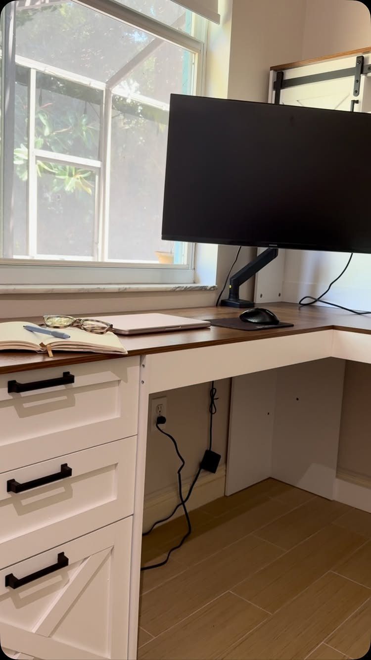 White corner desk with wooden top, black monitor, and drawers by a window
