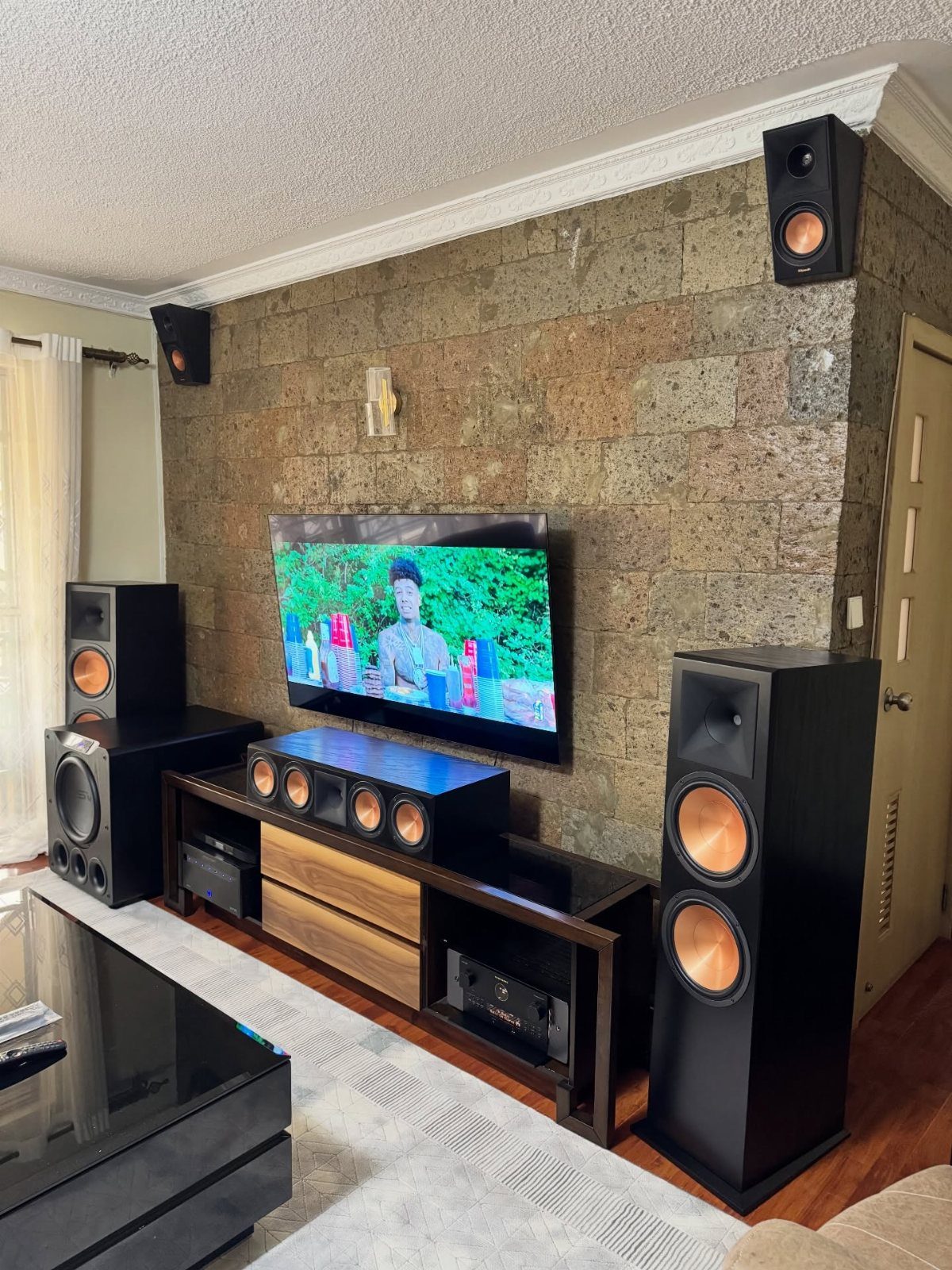 Wall-mounted television and surround sound speakers against a textured stone wall in a room.