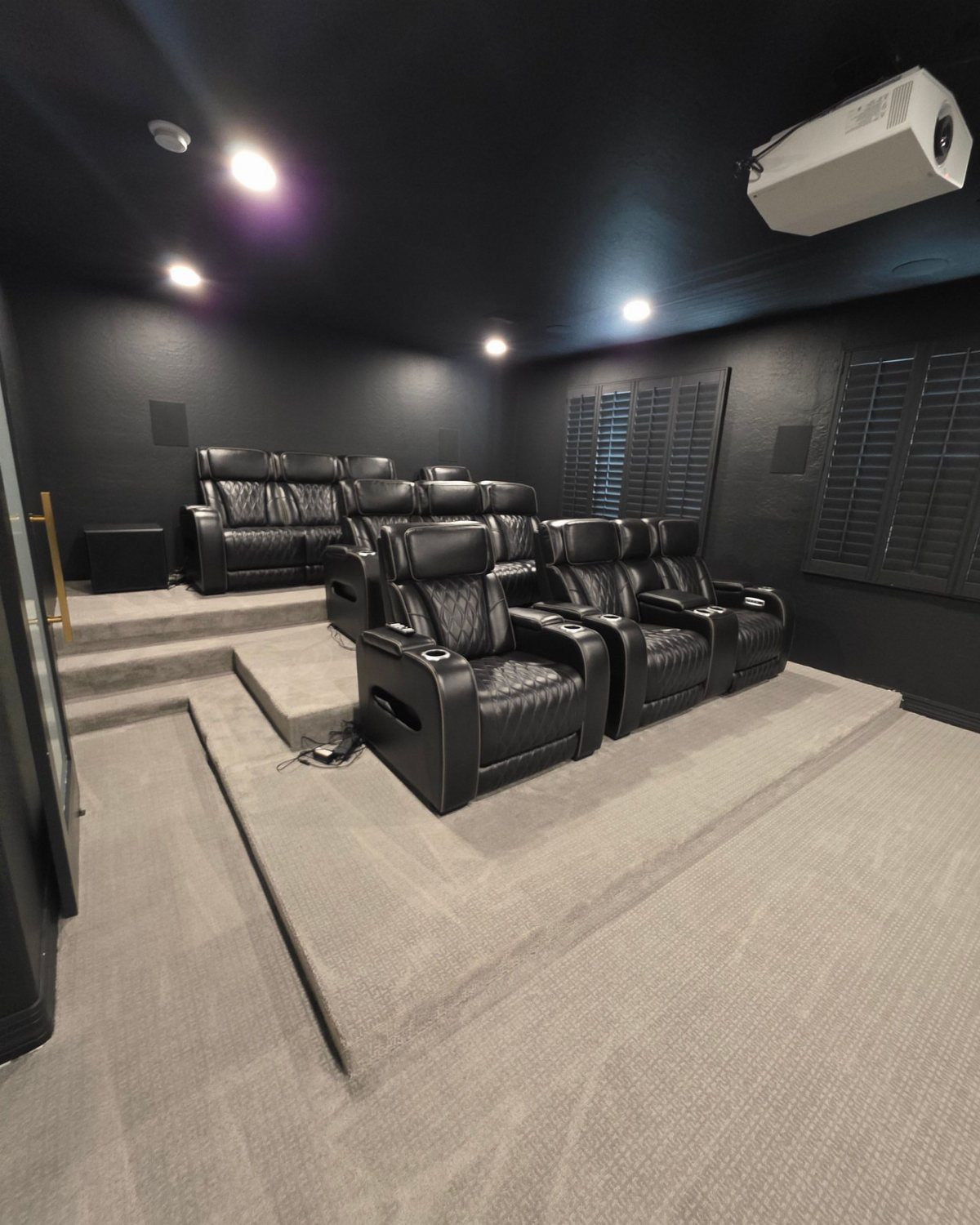 Black leather reclining cinema seats arranged on tiered grey carpeted platforms in a theater.