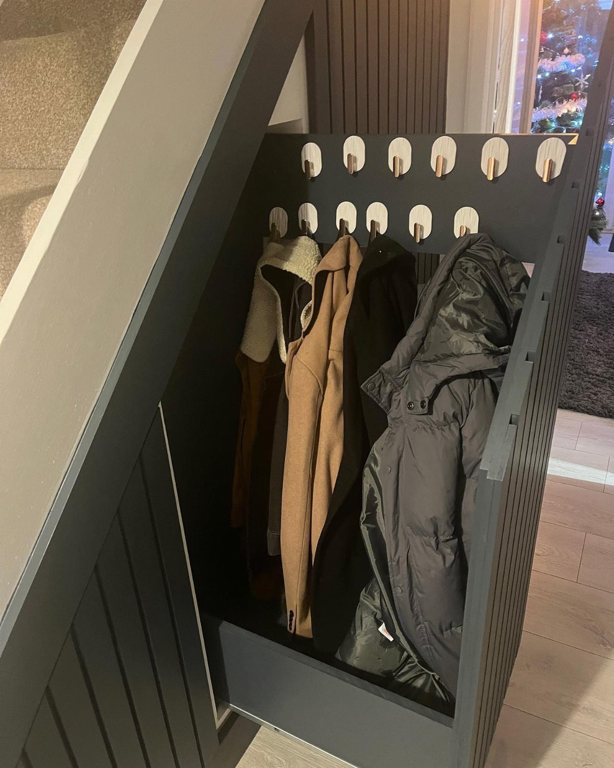 Dark grey built-in coat closet under stairs with winter jackets hanging on pegs.