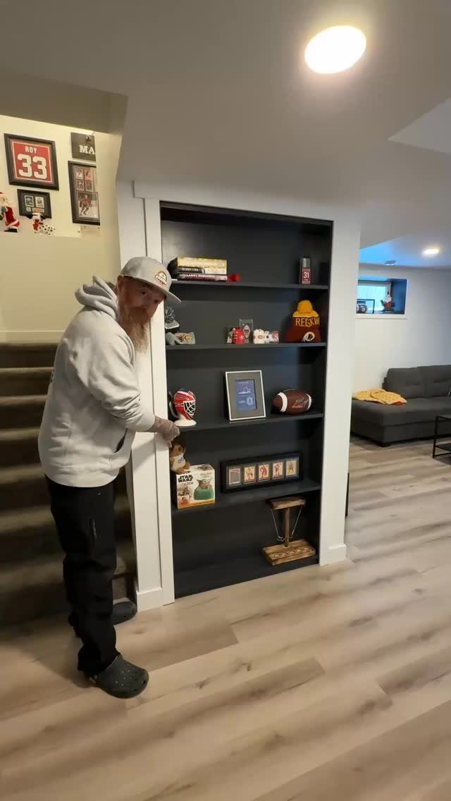 Built-in dark grey shelving unit under basement stairs displaying sports memorabilia and trophies.