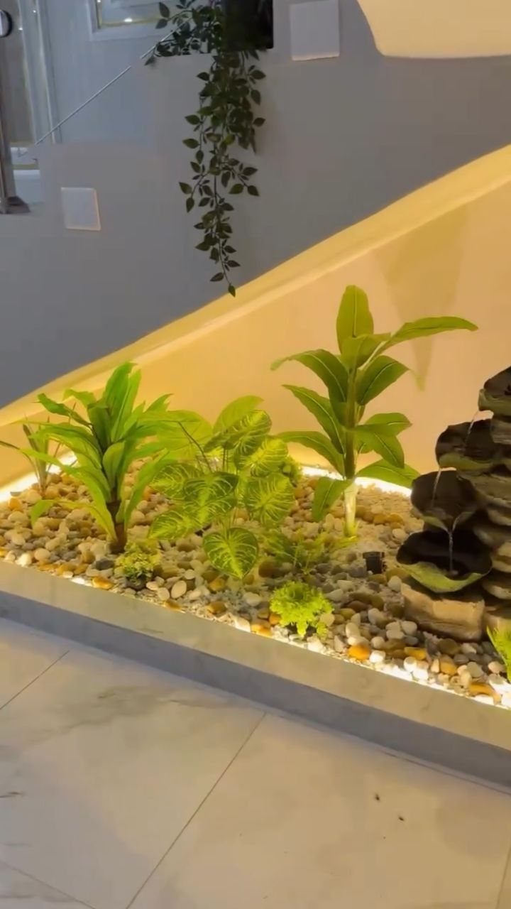 Indoor garden bed with green plants and pebbles under a white staircase.