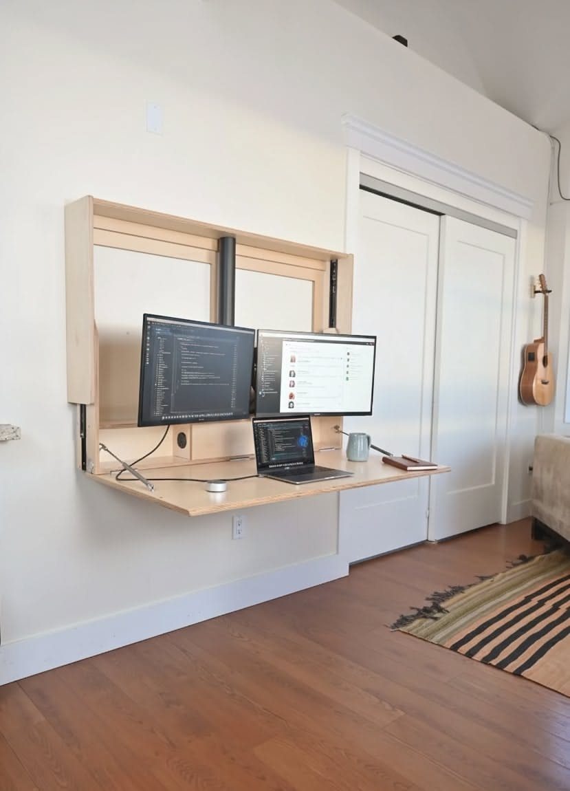Wall-mounted folding wooden desk with two monitors and a laptop in a white room.