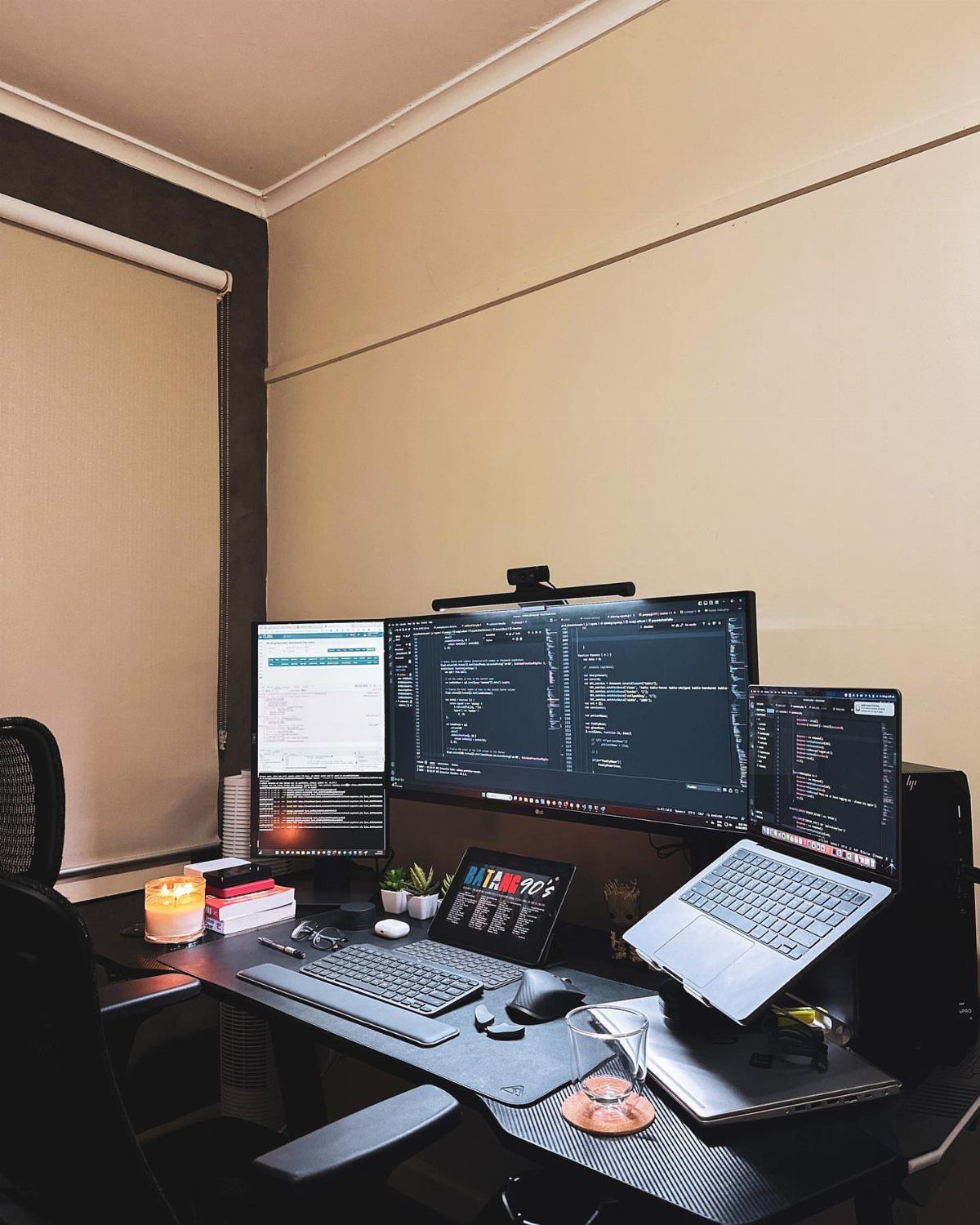 L-shaped desk with three monitors displaying code, a laptop, and office accessories.