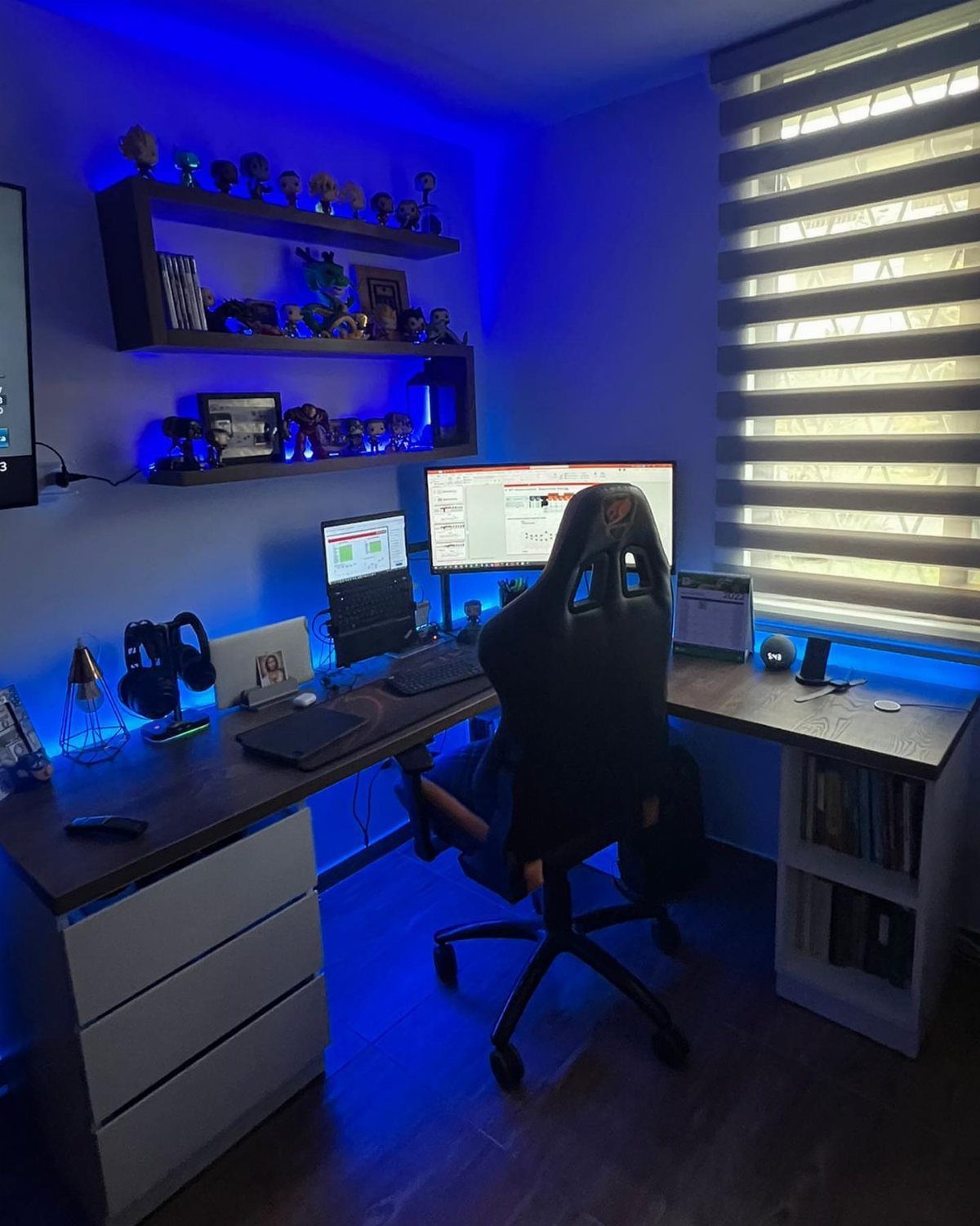 L-shaped desk with dual monitors, gaming chair, and blue LED lighting in a room.