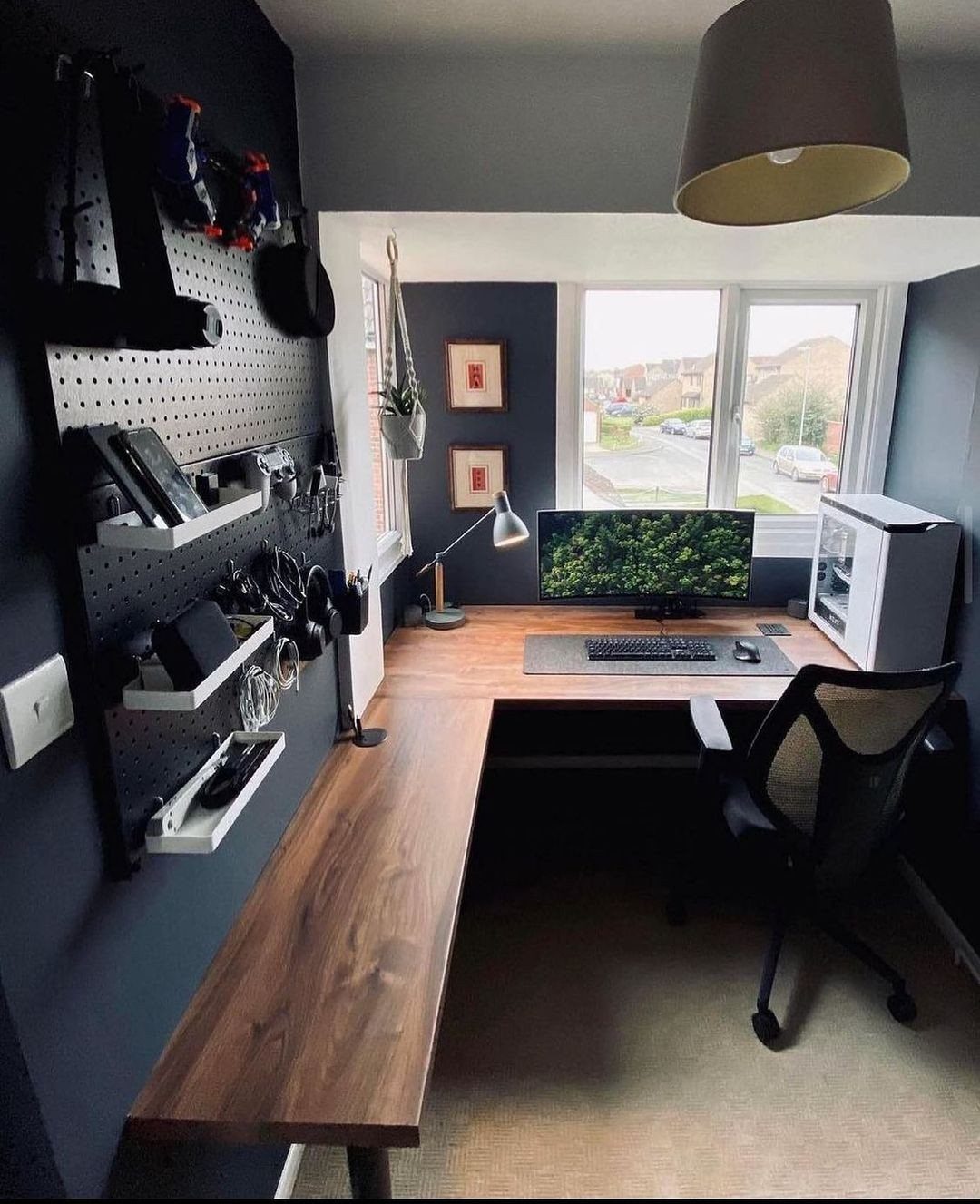 L-shaped wooden desk with a monitor, PC, and black pegboard on the wall