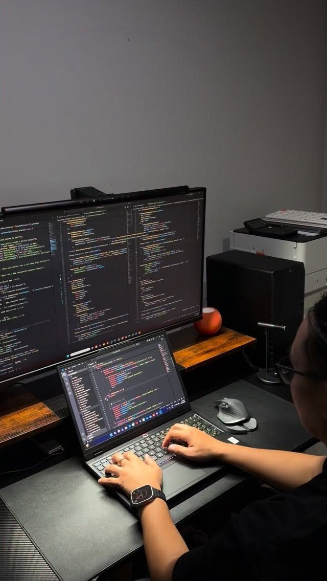 Person using a laptop and large monitor displaying code on a black desk