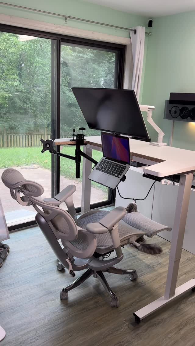 White standing desk with a large monitor, laptop on mount, and ergonomic chair.