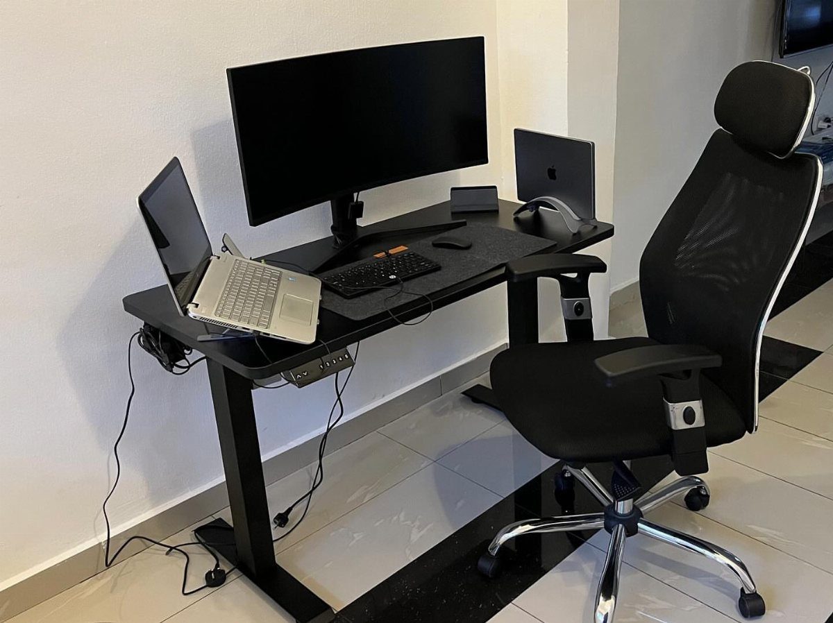 Black standing desk with a large monitor, laptop, and ergonomic office chair.
