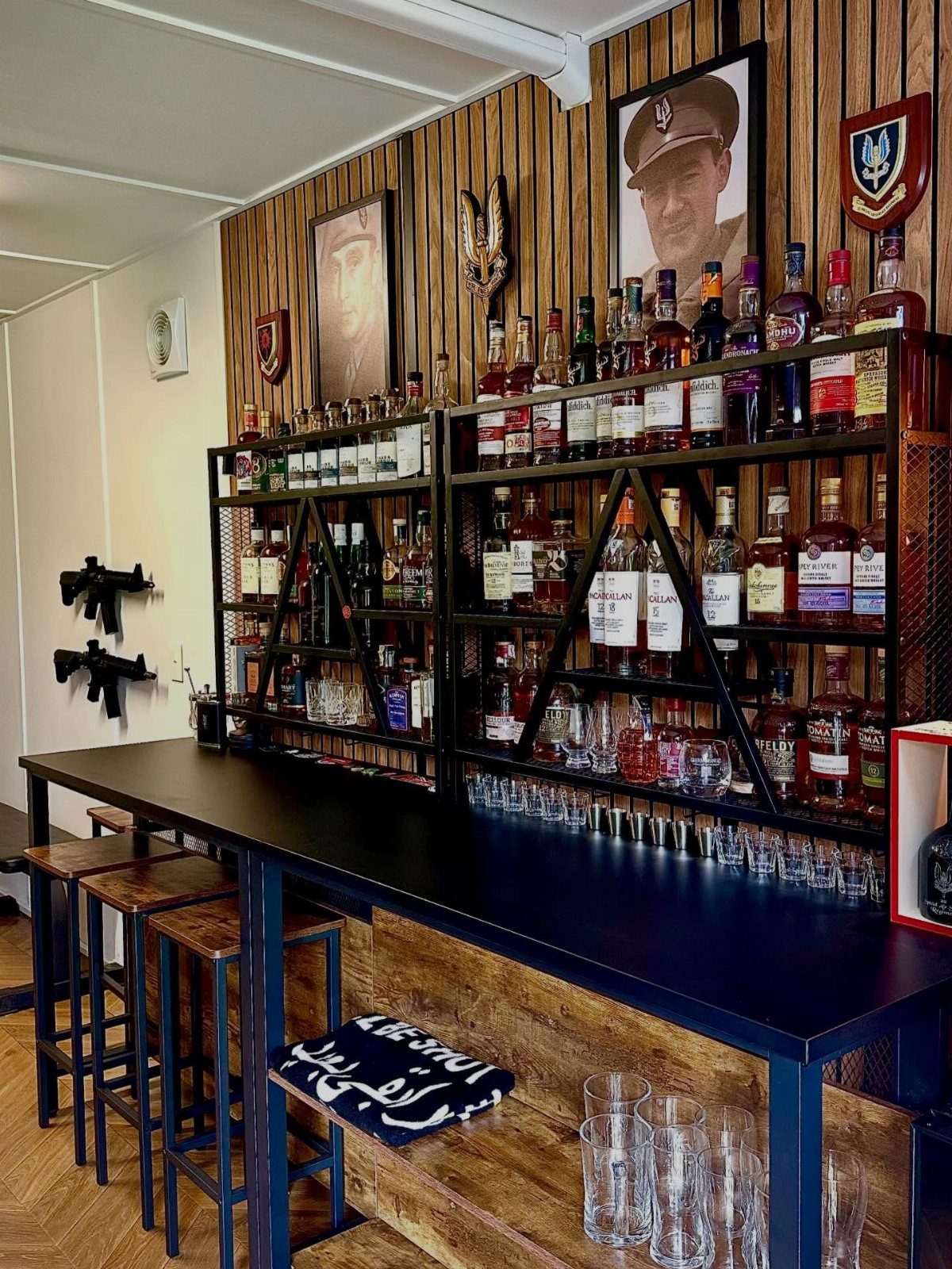 Home bar with black countertop, wooden stools, and liquor bottles on metal shelves.