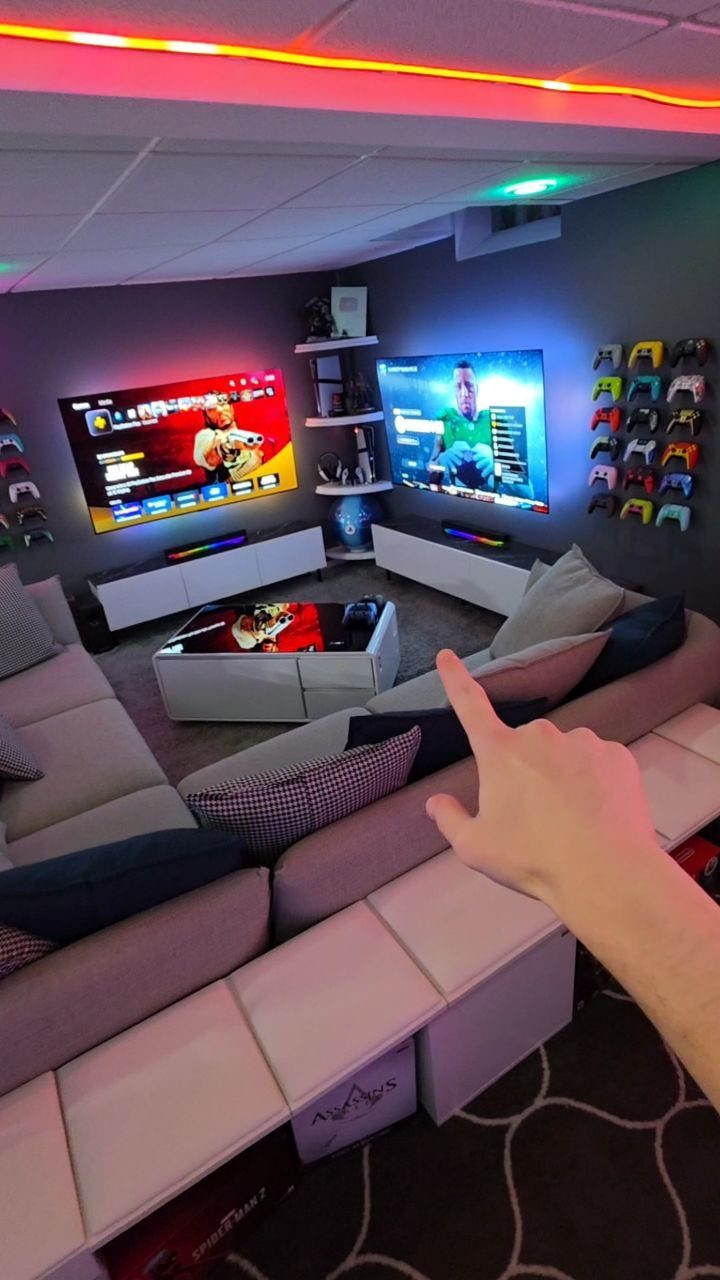 Modern gaming room with two large screens, grey sectional sofa, and controller wall.