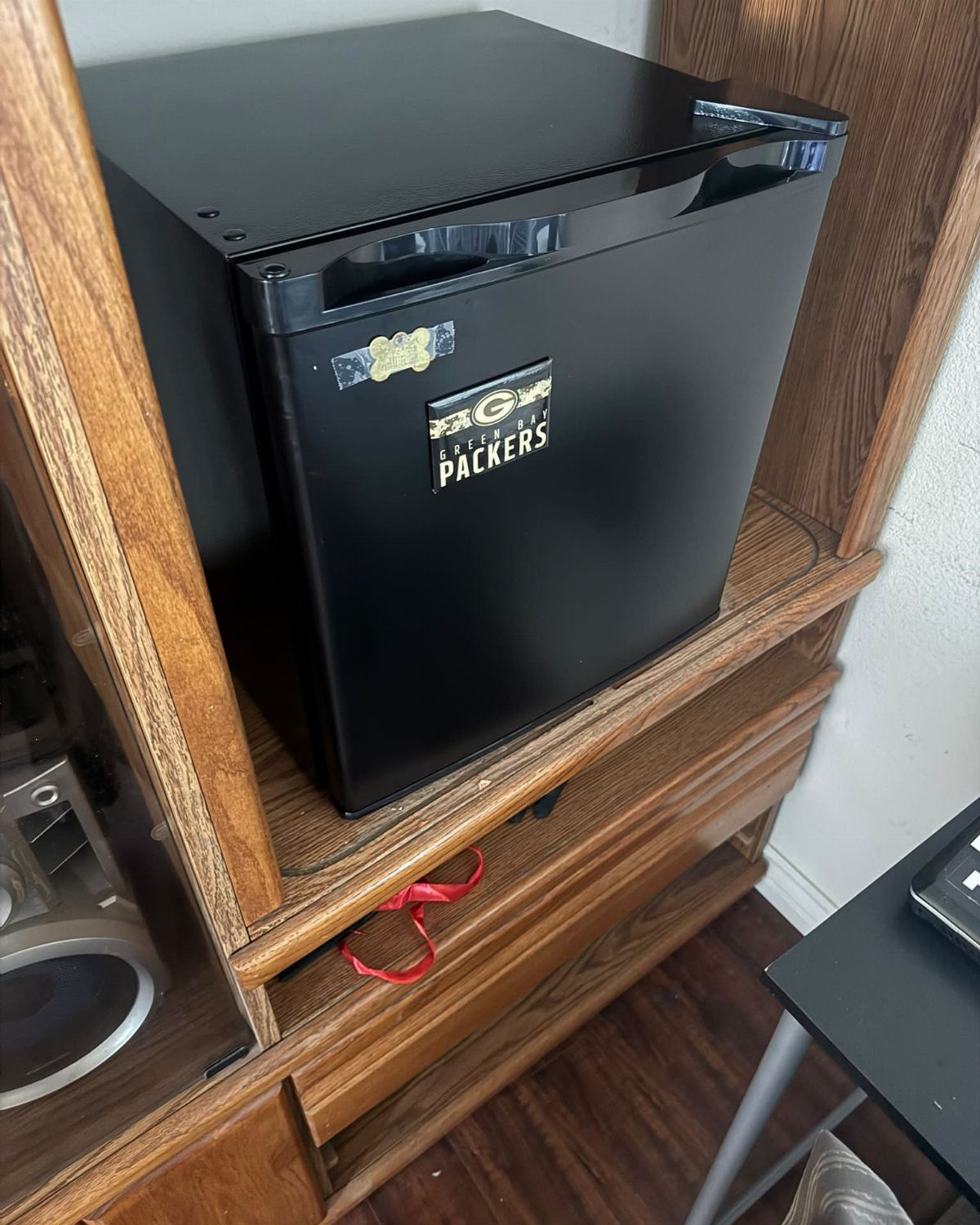 Black mini fridge with Packers sticker inside a wooden entertainment center shelf