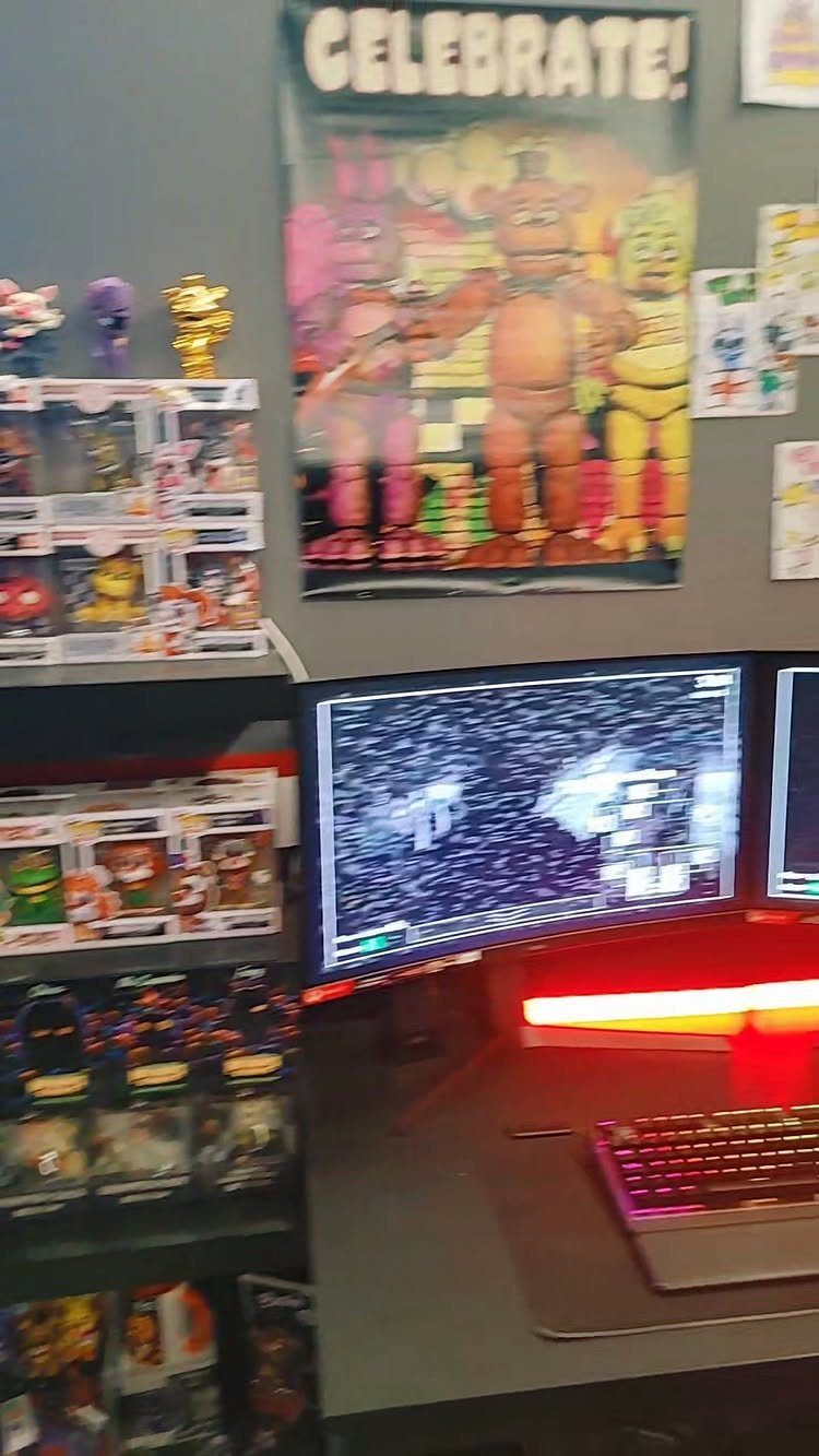 Gaming desk with dual monitors, RGB keyboard, and game-themed posters and collectibles.