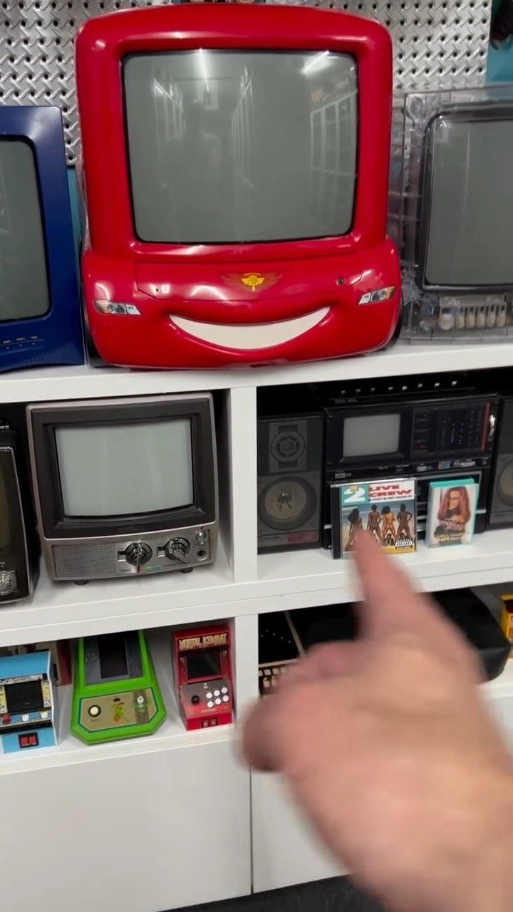 White modular shelving unit displaying vintage televisions, a stereo, and handheld gaming consoles.