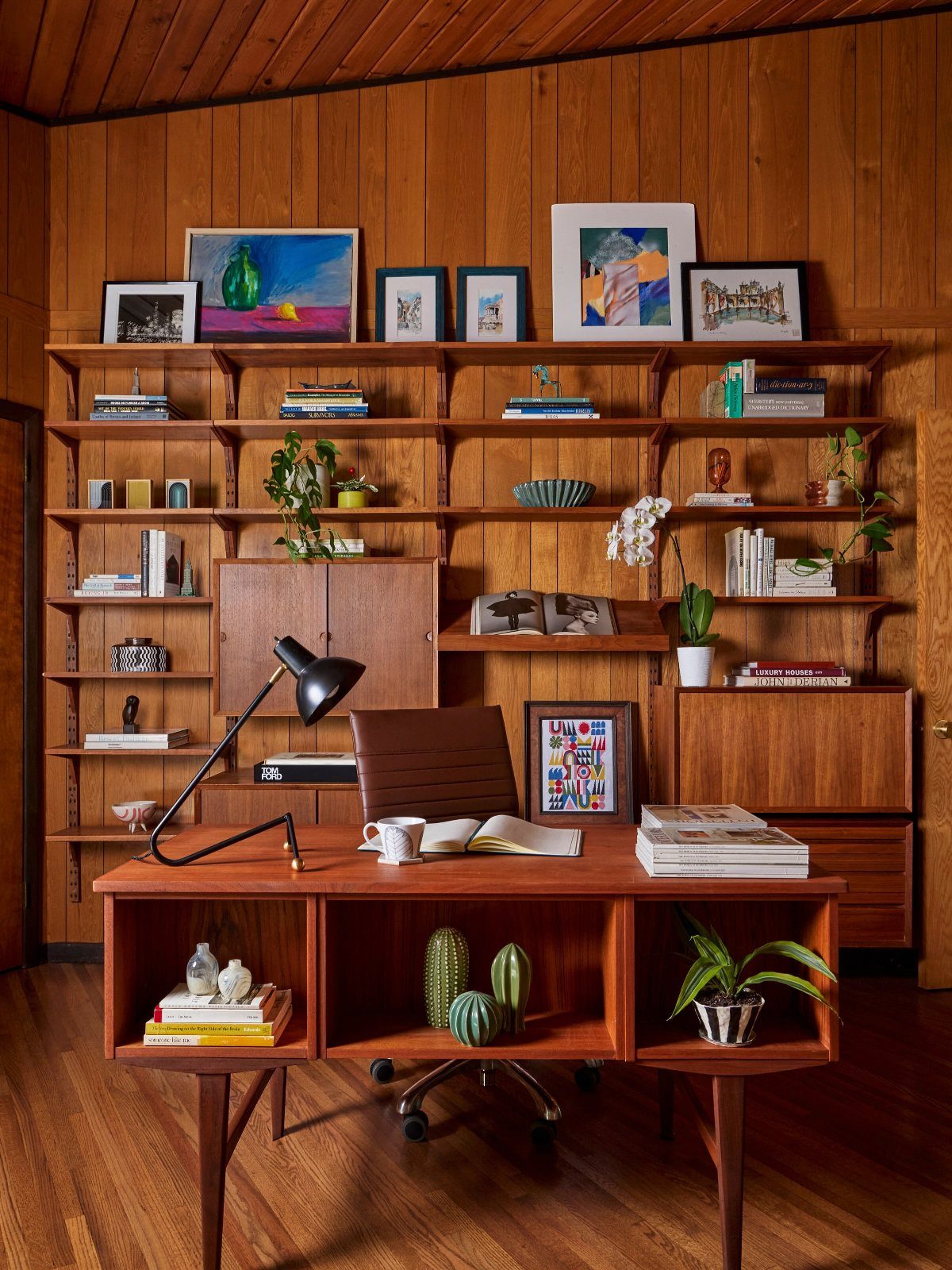Mid-century modern office with wooden desk, leather chair, and extensive wall shelving unit.