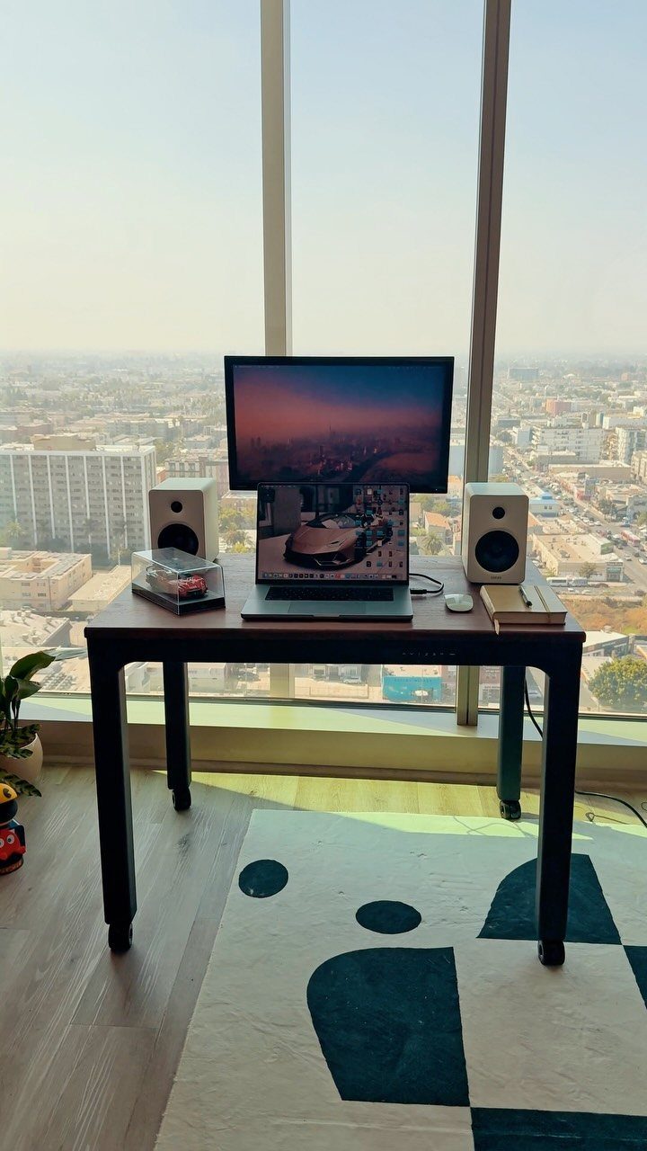 Dark wood desk with computer equipment placed before a large city view window.