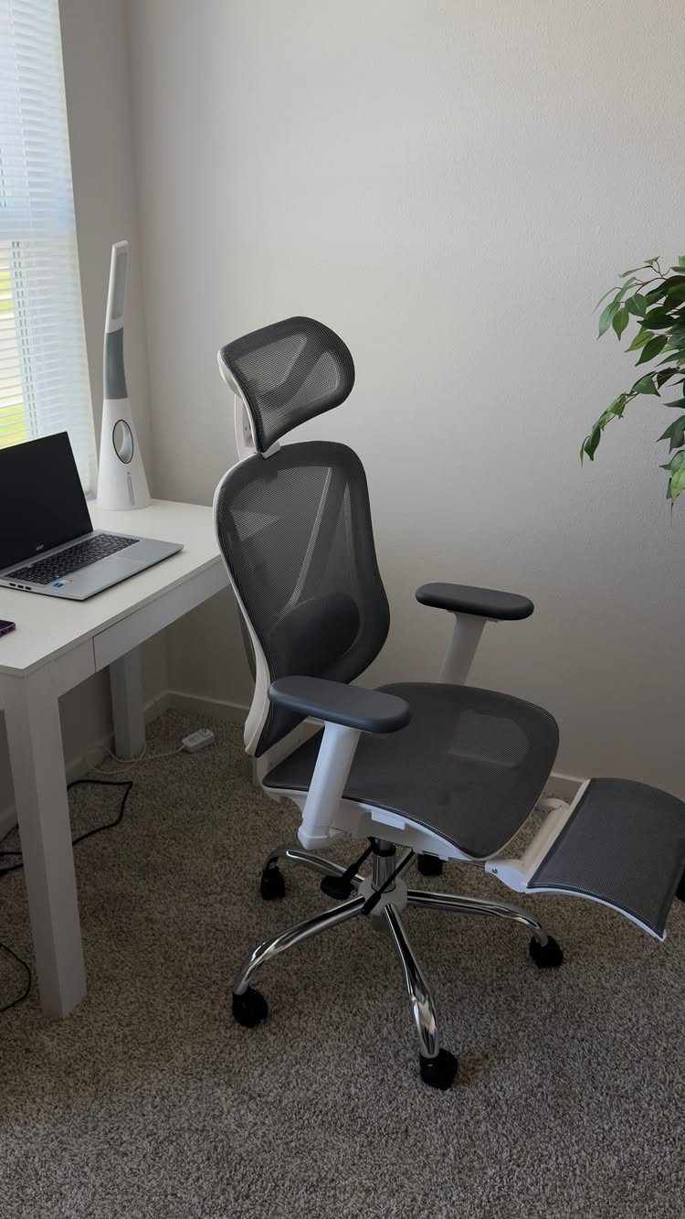 Ergonomic mesh office chair with headrest and footrest next to a white desk.