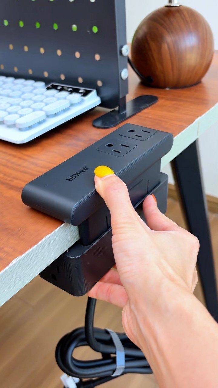 Black Anker power strip clamped to a wooden desk with a hand touching it.
