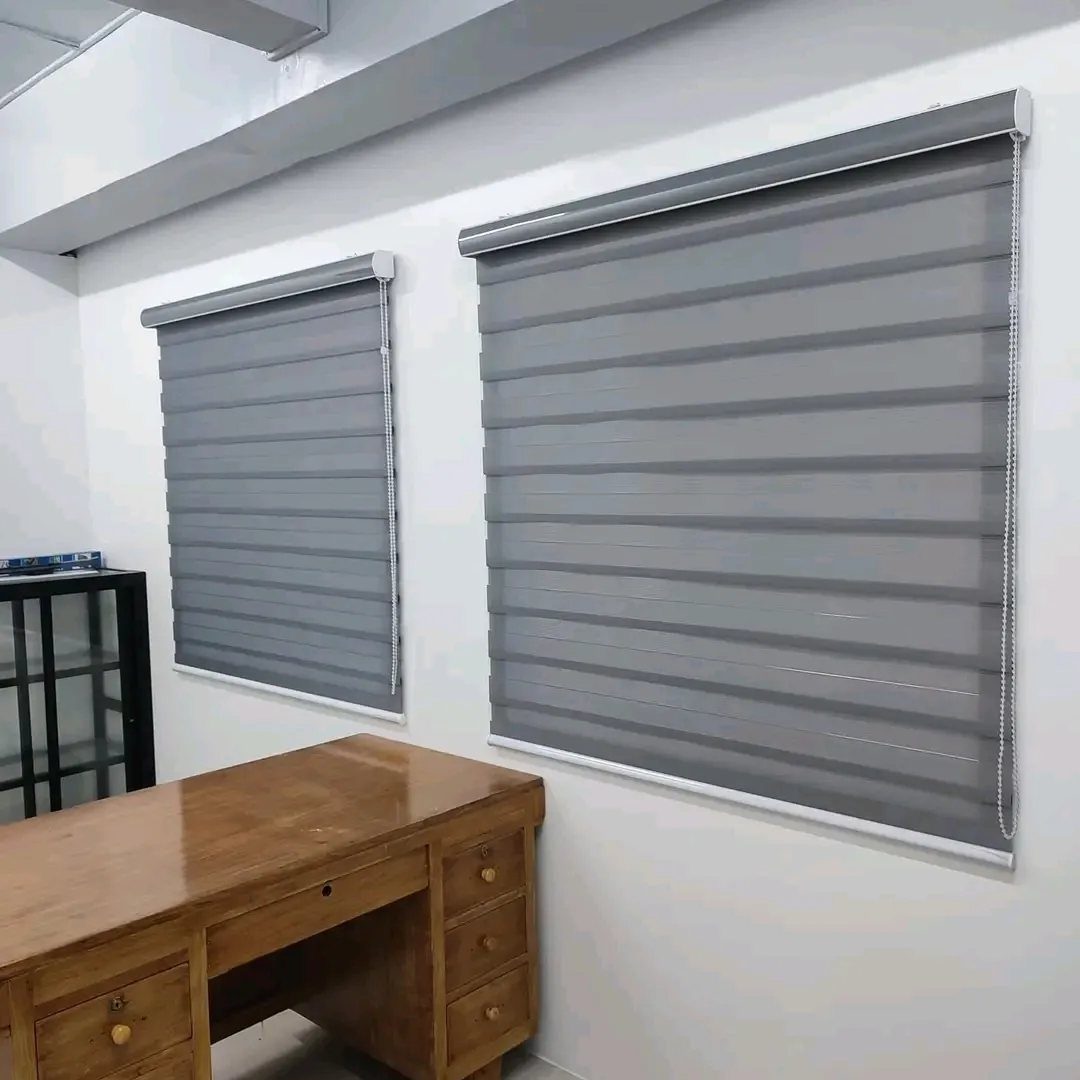 Grey zebra blinds installed on white walls above a wooden office desk.