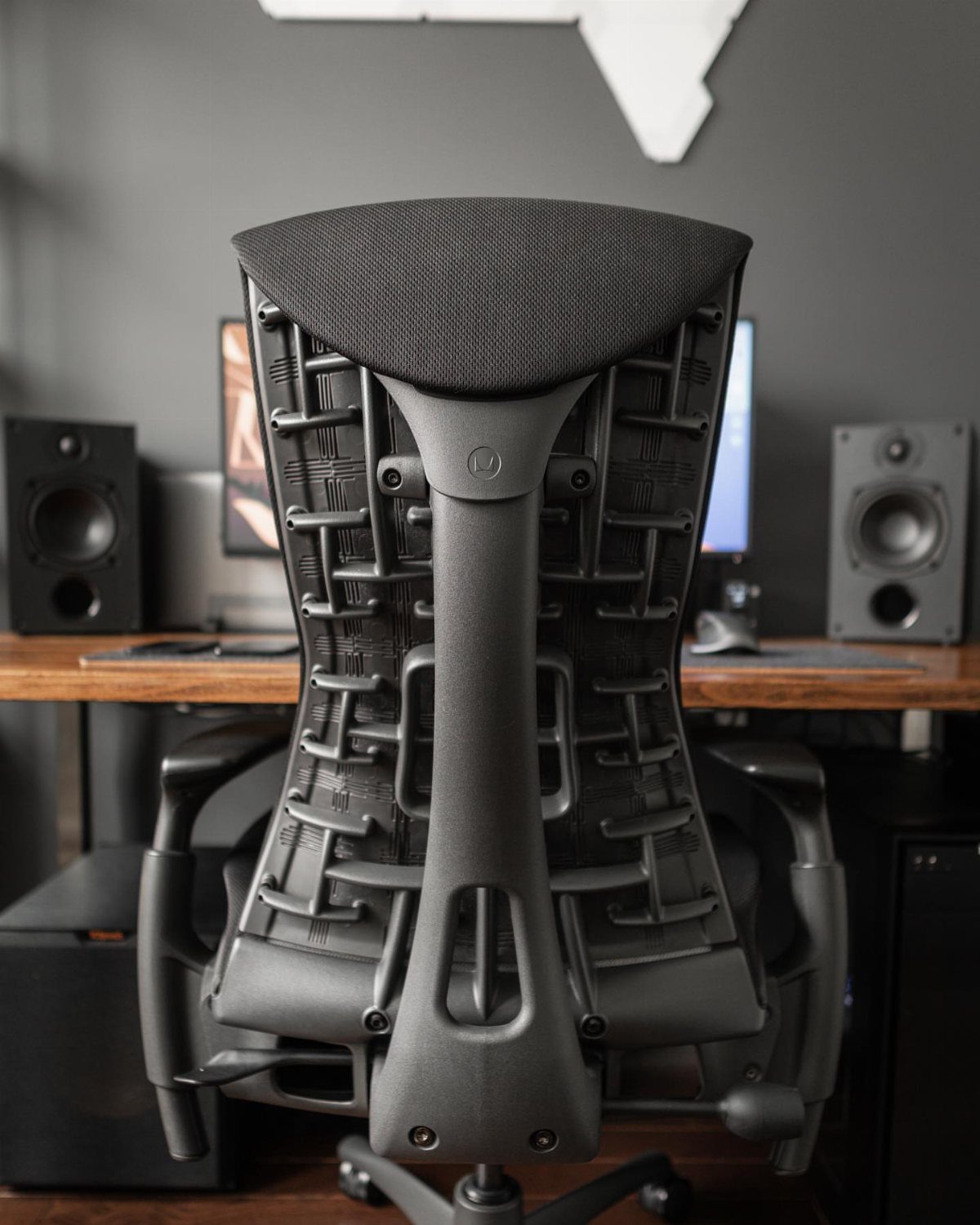 Grey ergonomic office chair with mesh back facing a wooden desk and speakers.