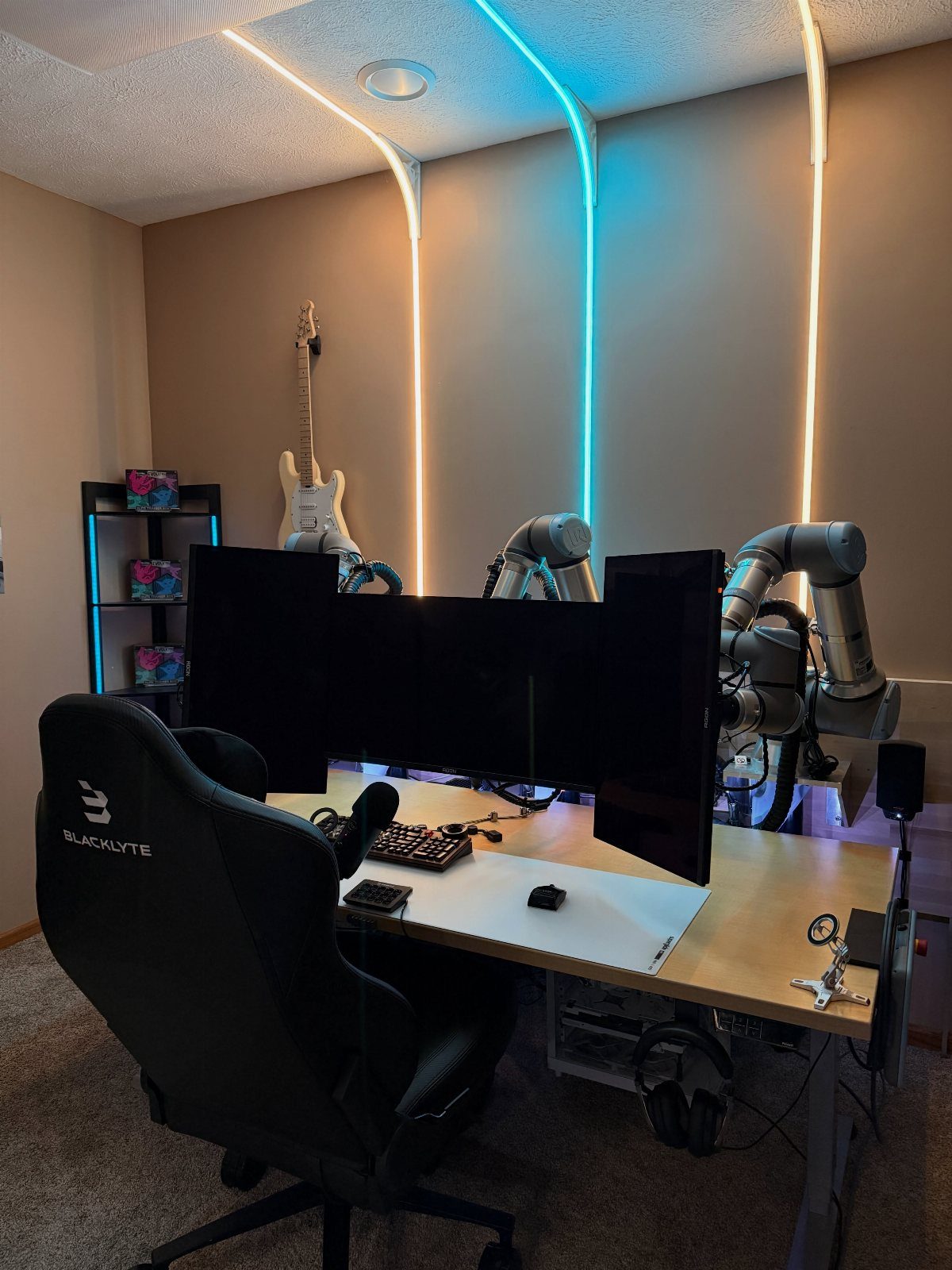 Gaming desk with triple monitors, black chair, and blue and white LED wall lighting.