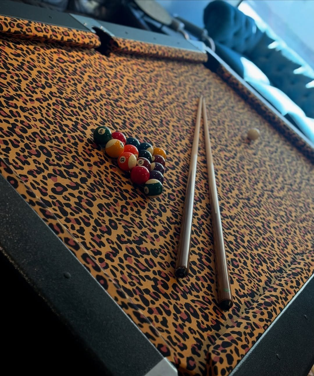 Leopard print pool table with colorful billiard balls and two wooden cues.
