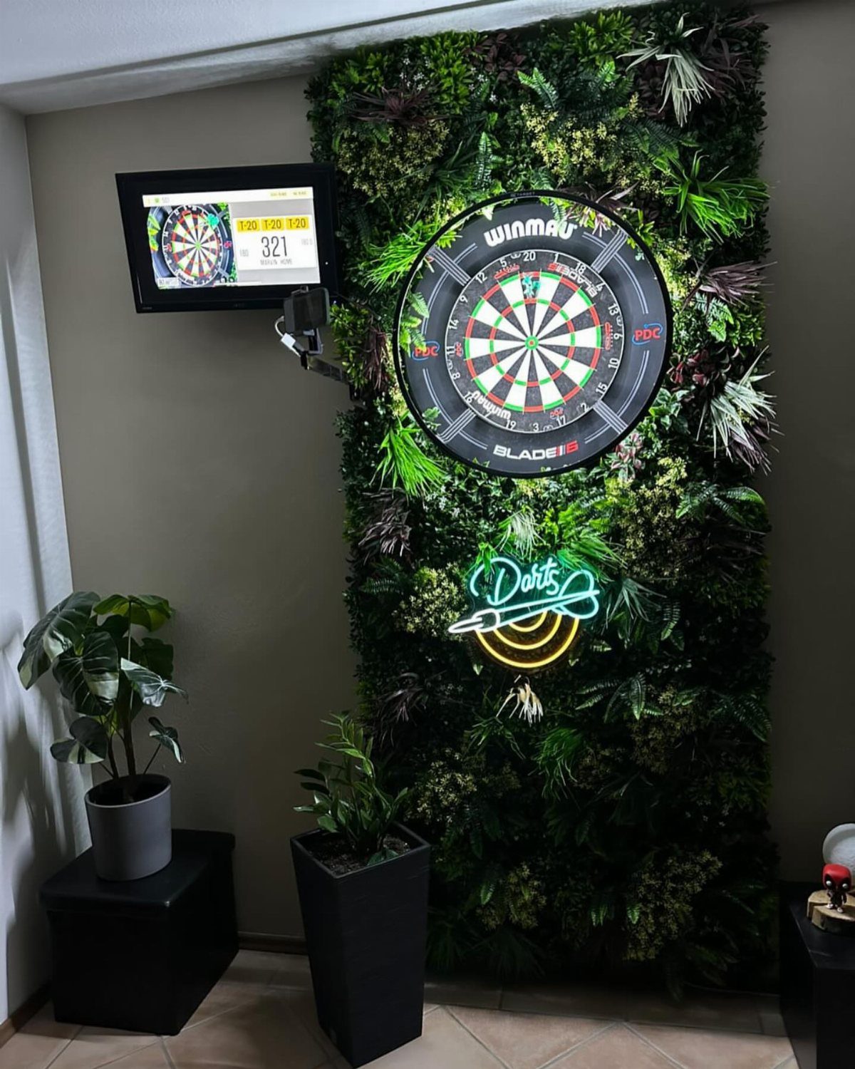 Dartboard mounted on a green plant wall with a neon sign and scoring screen.