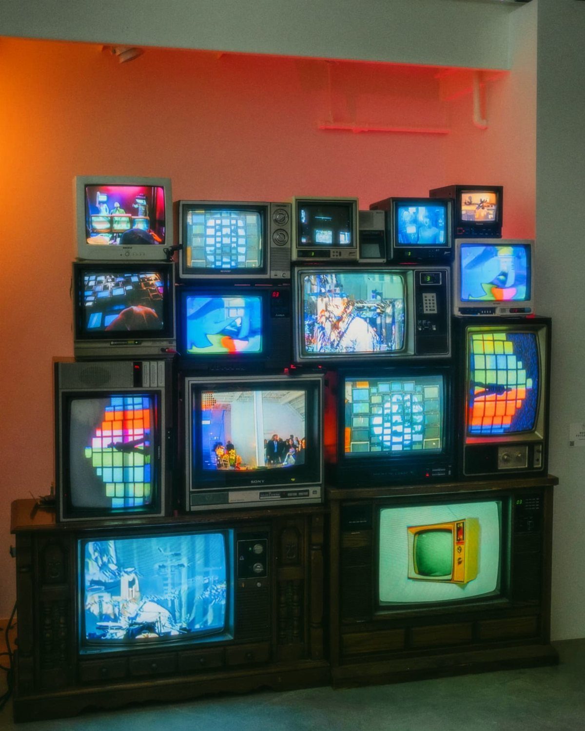 Stacked vintage CRT televisions displaying colorful images against a light wall with red lighting.
