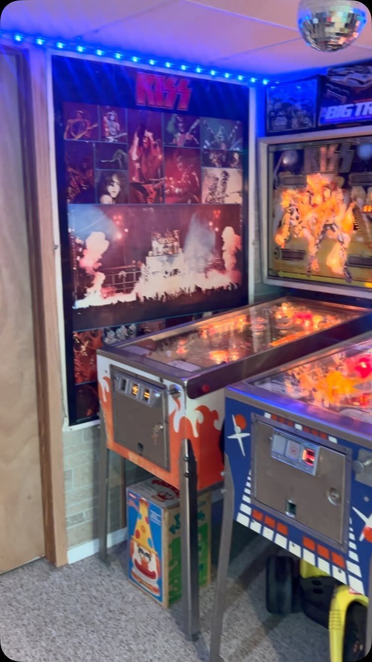 Two vintage pinball machines in a room with a KISS poster and LED lighting.