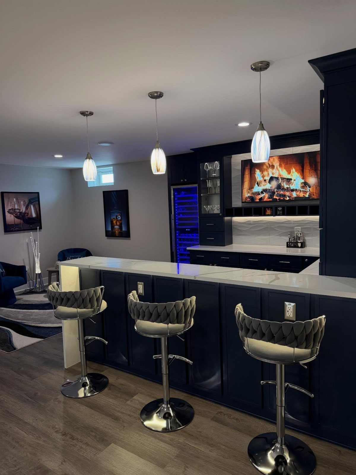 Modern basement bar with white countertops, dark blue cabinets, and grey quilted stools.