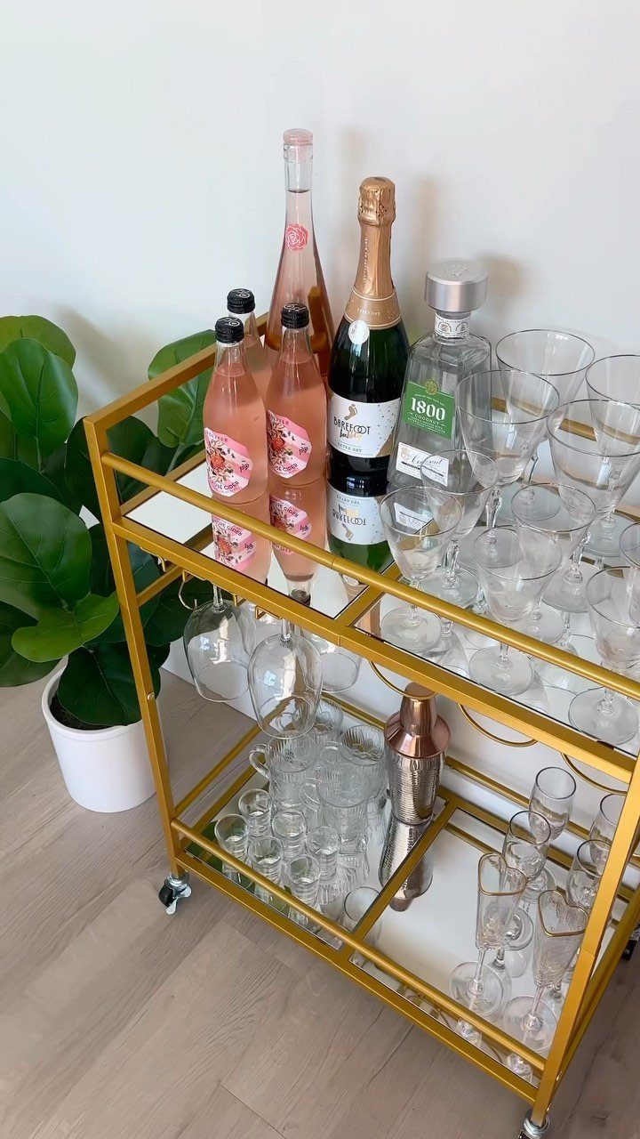 Gold bar cart with white shelves holding wine bottles and glassware next to a plant