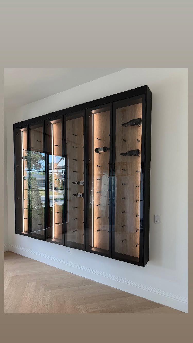 Wall-mounted glass wine cellar with black frame, wooden shelves, and warm interior lighting.