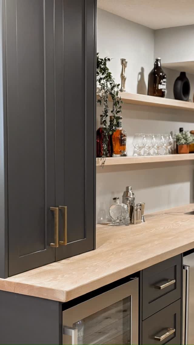 Dark grey home bar with light wood countertops and open shelving for glassware.