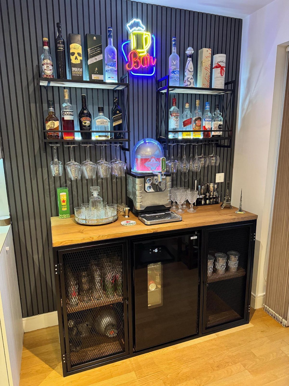 Black metal home bar with wooden top, liquor bottles, and built-in refrigerator.