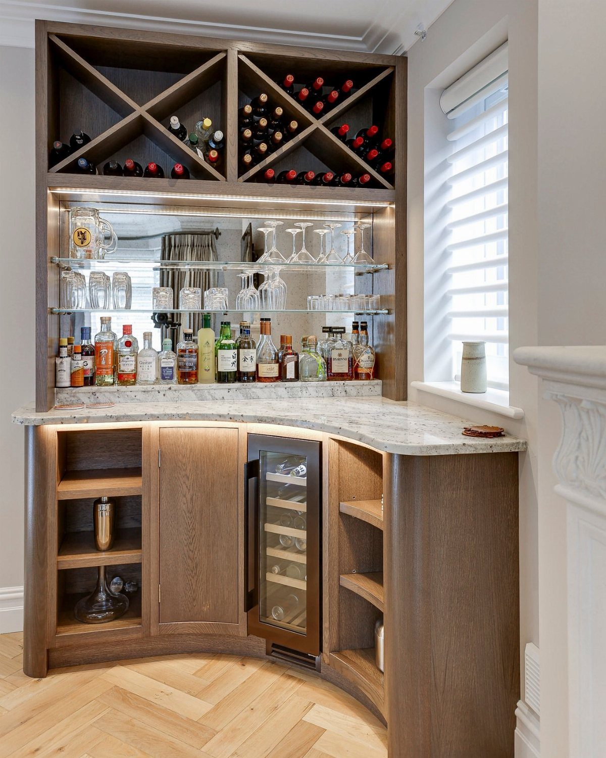 Curved wooden home bar with marble top, wine refrigerator, and built-in wine rack.