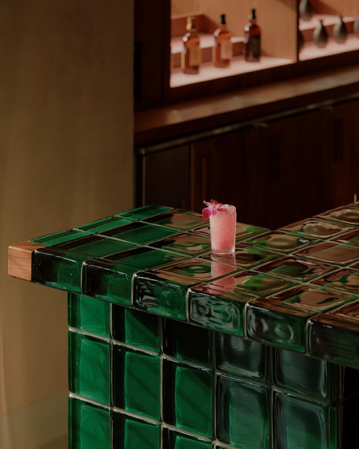 Green glass block home bar counter with a pink cocktail and liquor bottles.