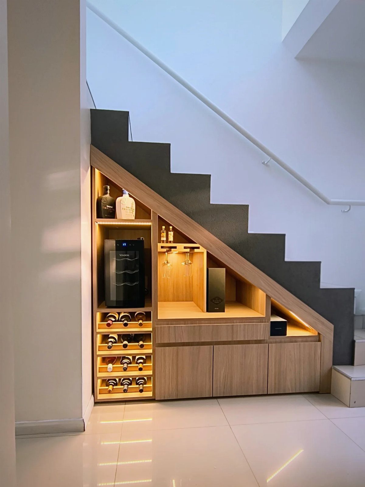 Wooden wine storage unit with cooler and bottle racks under a grey staircase.