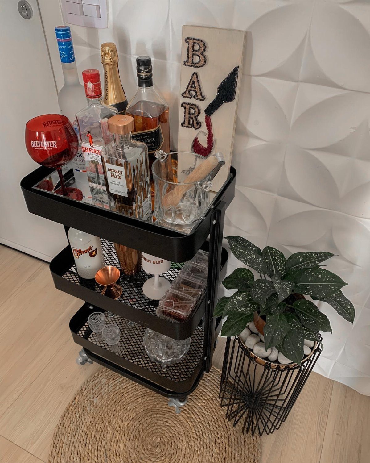 Black three-tier bar cart stocked with liquor bottles and glasses next to a plant.