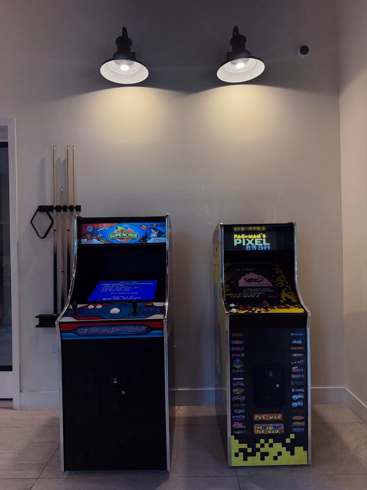 Two arcade game cabinets standing against a white wall with a pool cue rack.