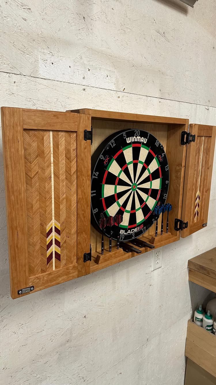 Wooden wall-mounted dartboard cabinet with open doors and a set of darts.