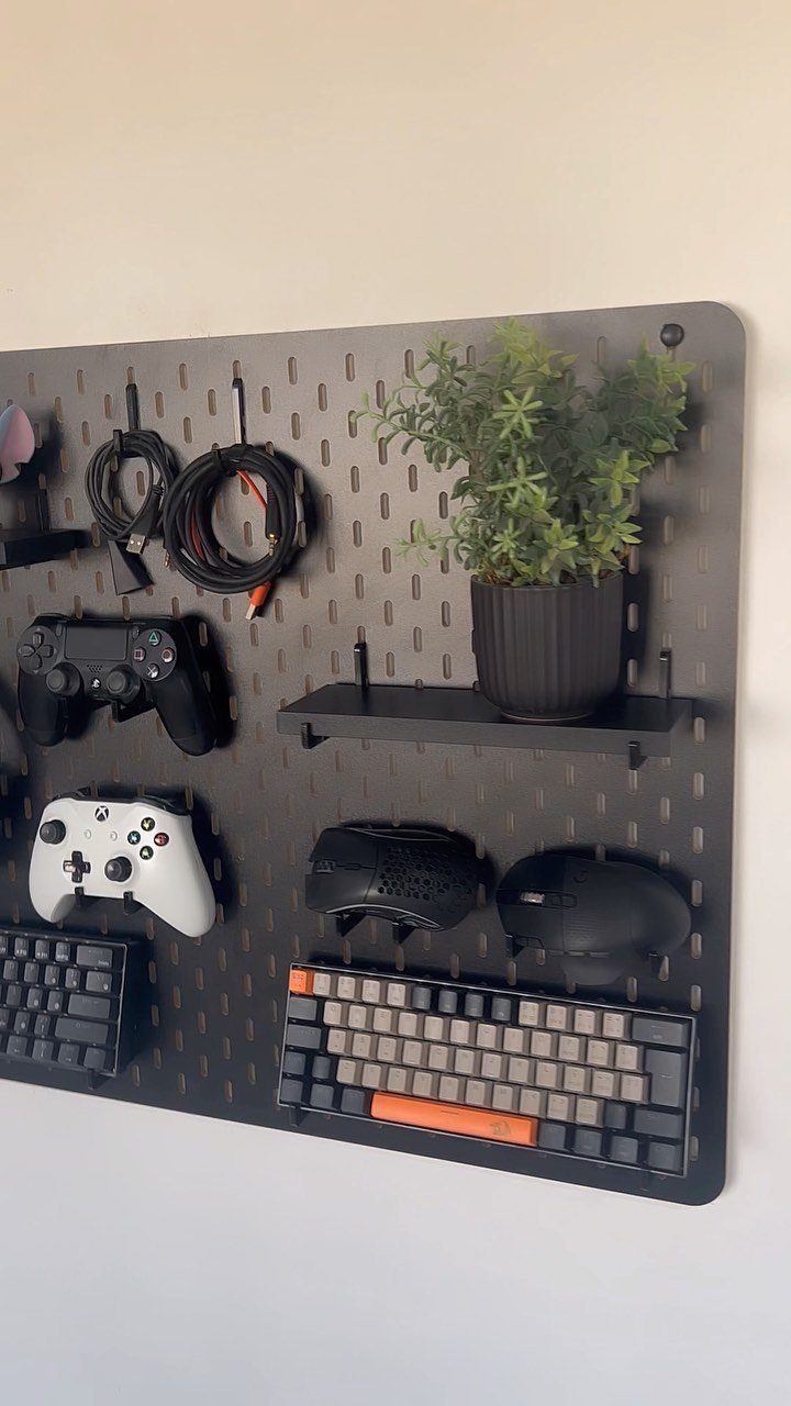Black pegboard organizing gaming controllers, keyboards, and mice with a small green plant.