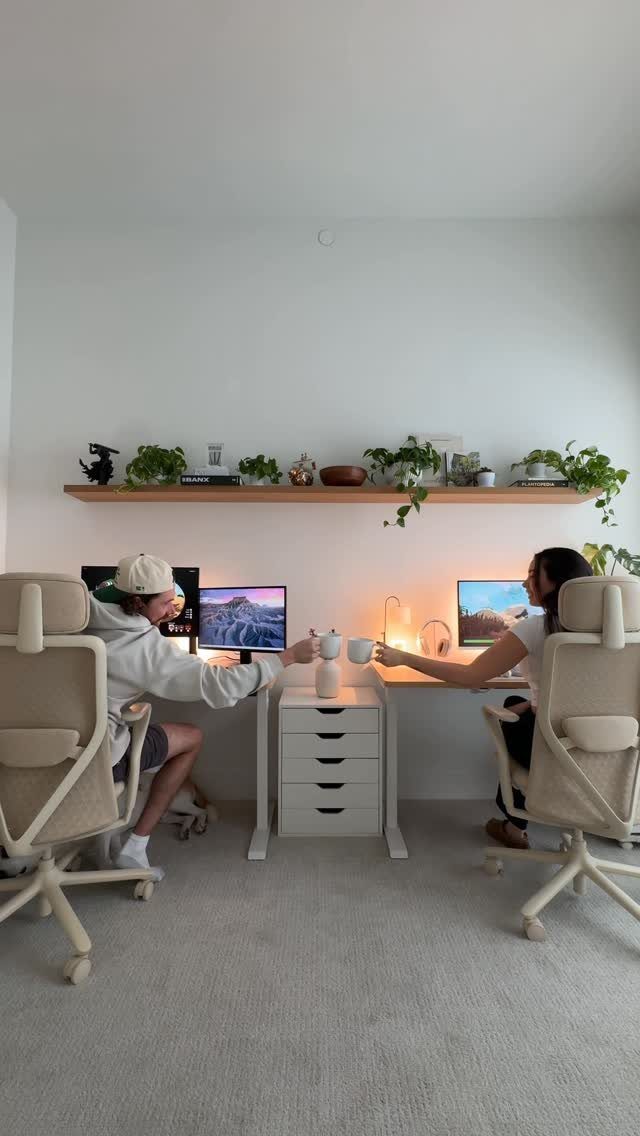 Two people working at a shared white desk with separate monitors and ergonomic chairs.