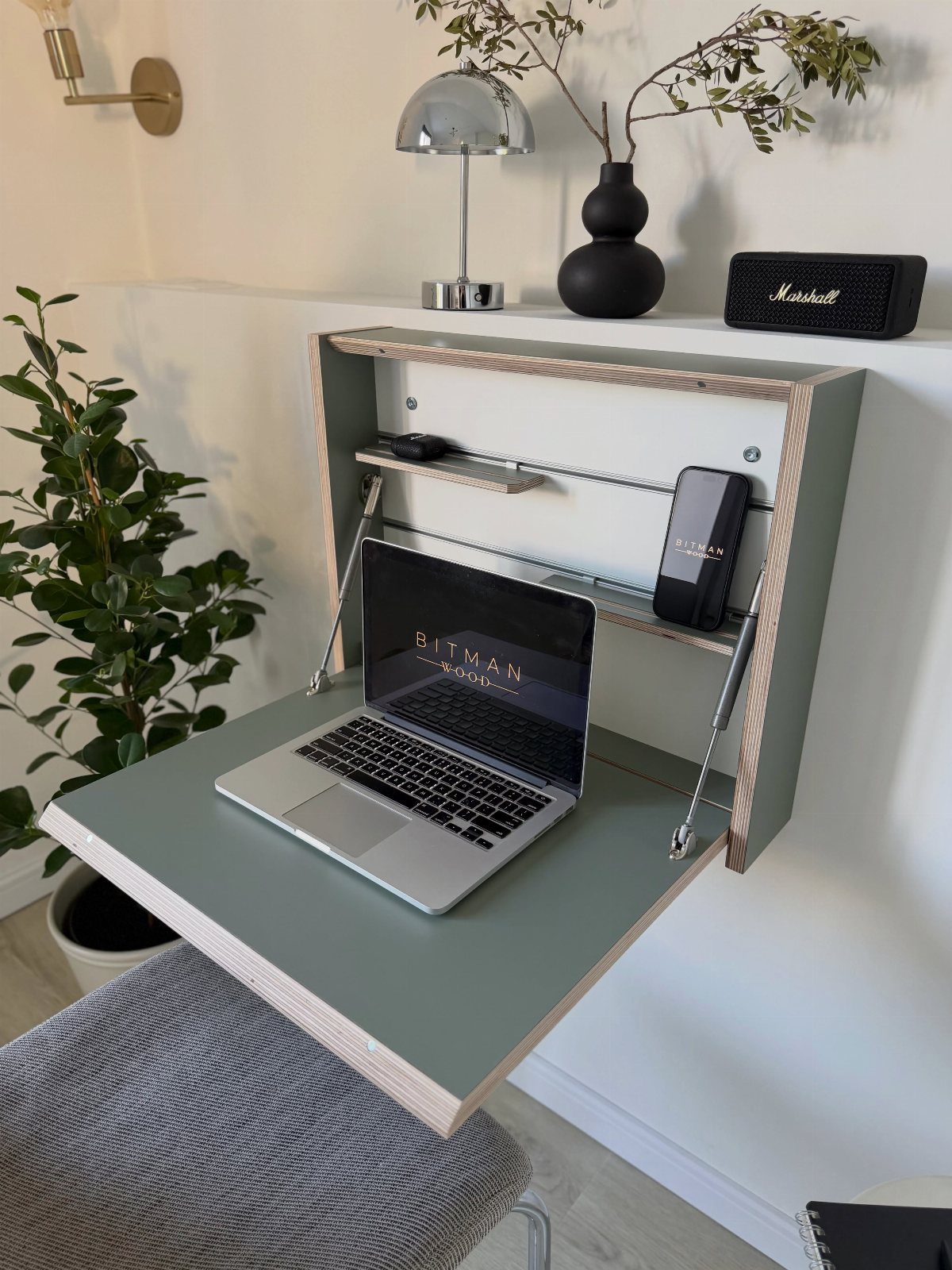 Wall-mounted folding sage green desk with integrated vertical shelving and a laptop.