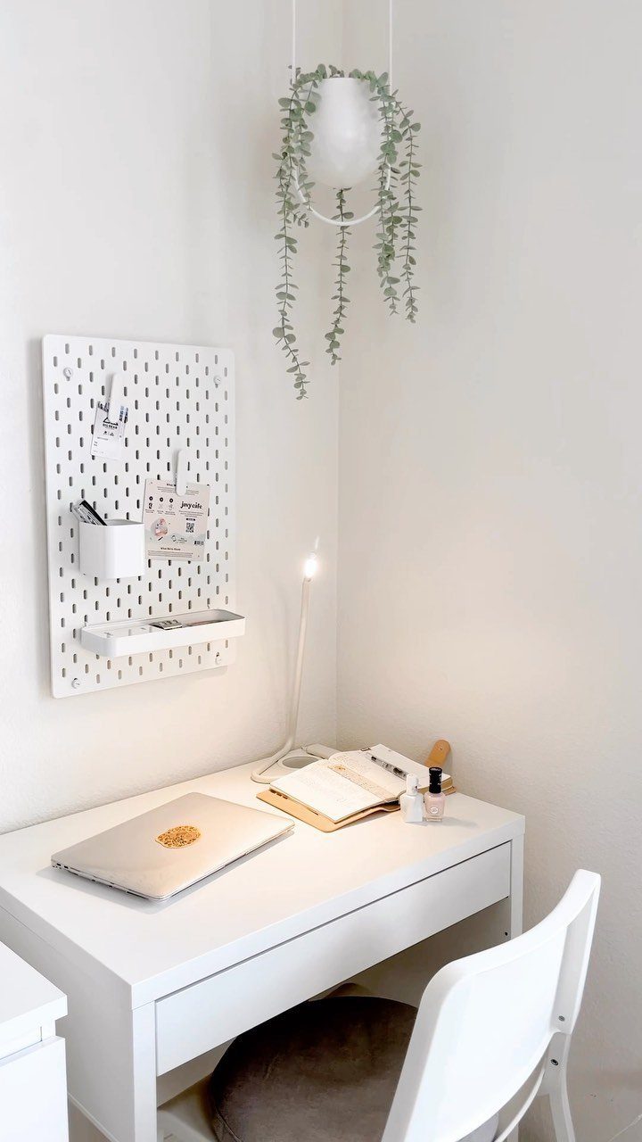 Small white writing desk with laptop, pegboard organizer, and hanging plant in white room