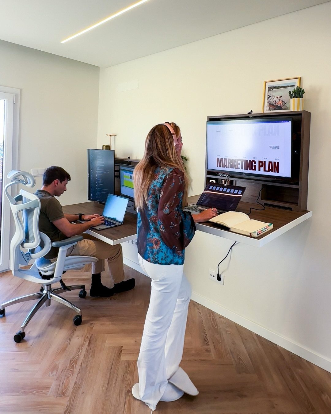 Two people working side by side at a long wall-mounted wooden workstation desk.