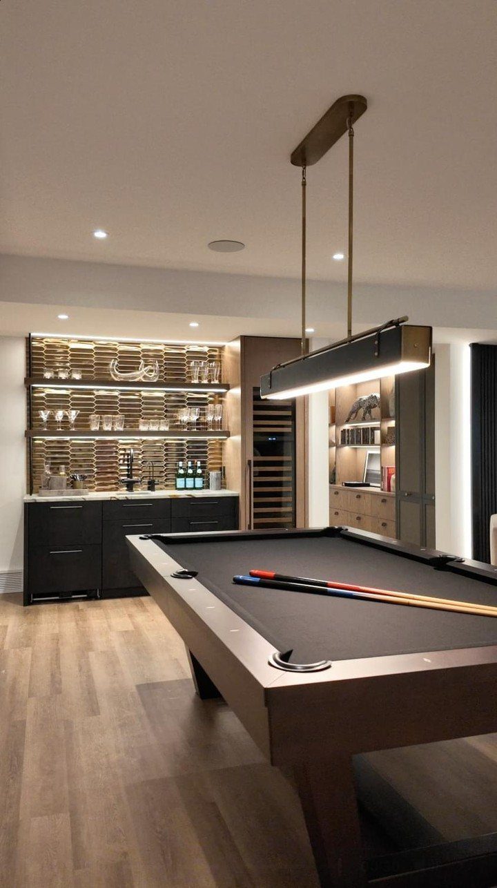 Modern pool table in a game room with a backlit bar and pendant lighting.